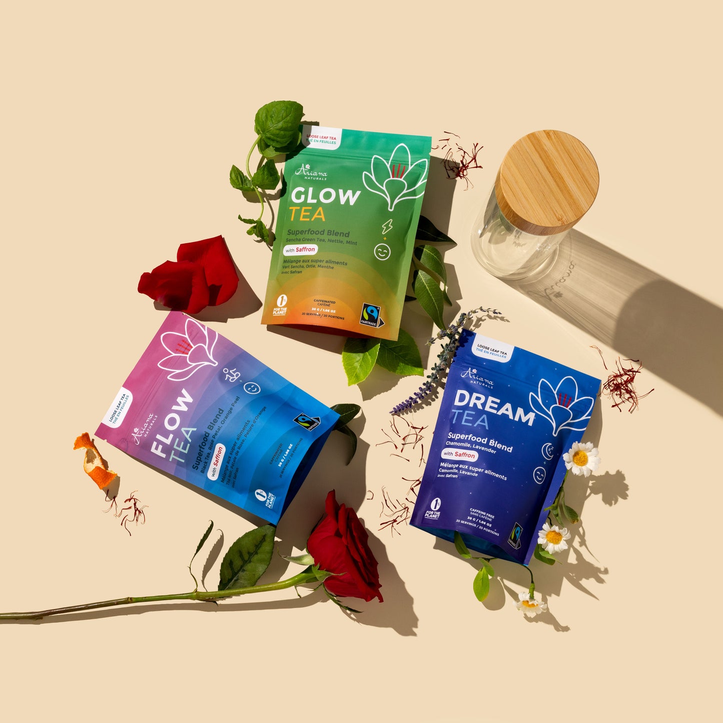 Three tea pouches from Ariana Naturals are arranged on a beige background with their ingredients, red roses, sencha green tea leaves, nettle leaves, black tea leaves, a mint sprig, lavender, chamomile, saffron, an orange twist and a glass bottle with a wooden lid. The pouches are labeled Glow Tea, Flow Tea, and Dream Tea.