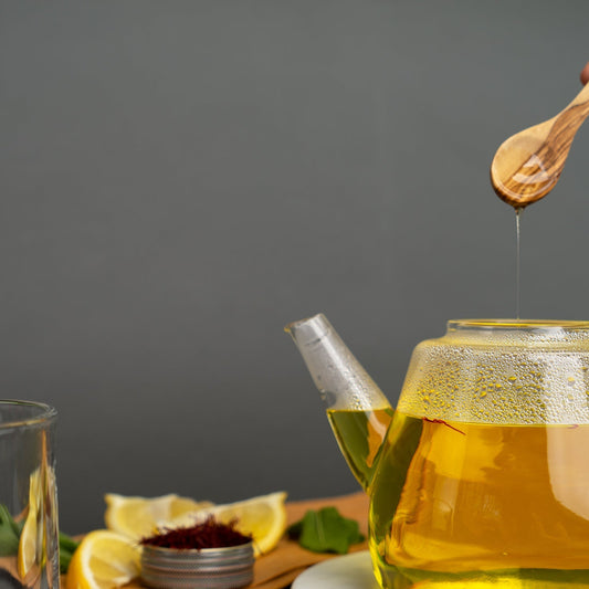 Saffron Tea in a tea pot with lemon and honey dripping into tea pot. A jar of saffron with lemons in the background. The background is grey.