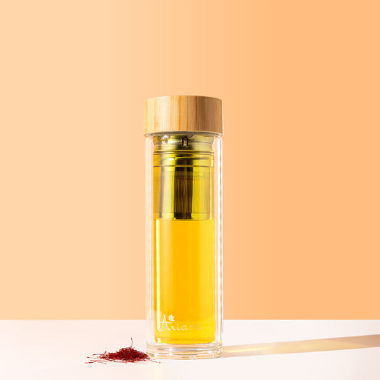 Clear glass bottle with wood lid containing a saffron tea, with a pile of saffron beside it on a white surface and peach background