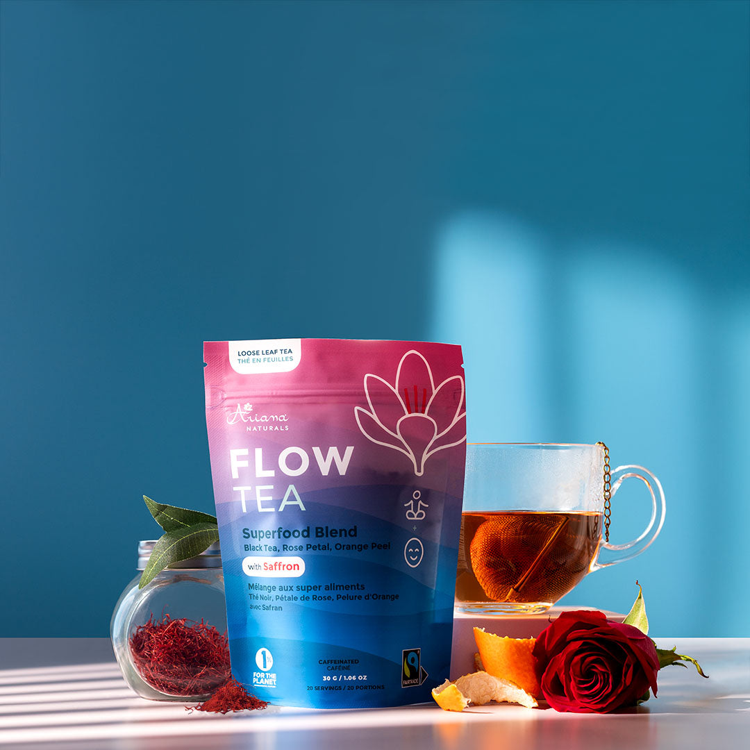 Ariana Naturals Flow Tea package with a cup of the tea and it's ingredients, a jar of saffron, black tea leaves, orange peel twists, and rose on a blue and light pink background. Morning light shining in.