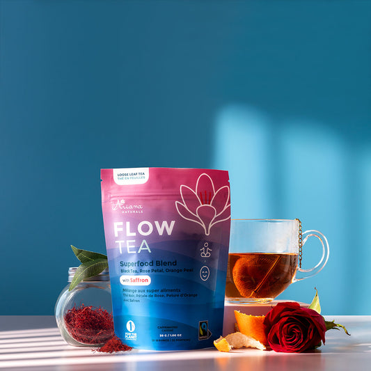 Ariana Naturals Flow Tea package with a cup of the tea and it's ingredients, a jar of saffron, black tea leaves, orange peel twists, and rose on a blue and light pink background. Morning light shining in.
