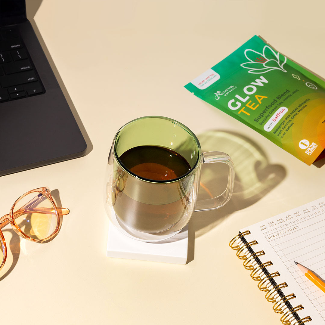 Green glass mug with tea, 'Glow Tea' package, blue light glasses, a notebook, and a laptop, on a pale yellow surface