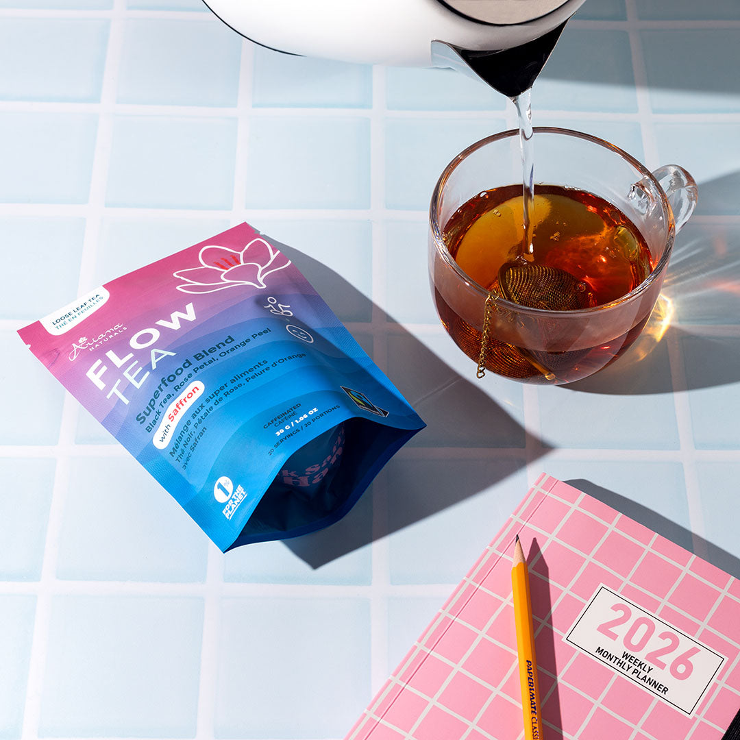 Water being poured into a glass with ball infuser full of  Ariana Naturals Flow Tea. With the package and a pink planner on a blue tiled surface.