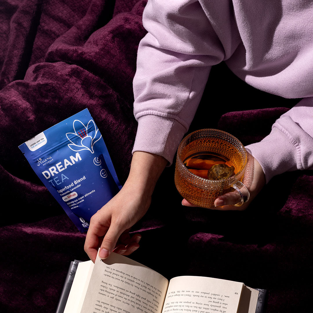 Person holding a cup of Ariana Naturals Dream tea and a book with a packet of Dream Tea on a purple blanket.