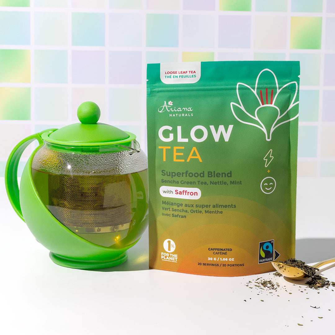 A stand-up pouch of Ariana Naturals Glow Tea Superfood Blend with Safrron, shown next to a green teapot filled with the tea and a small gold spoon with loose tea, against a pastel tiled background and white surface.