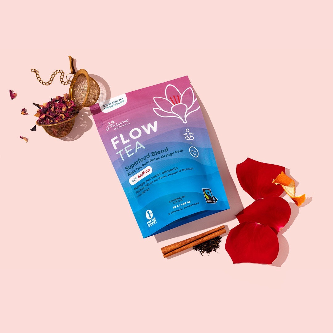 A package of Ariana Naturals Flow Tea Superfood Blend is displayed on a light pink background, styled with dried flower petals in a small ball tea infuser, cinnamon sticks, and red rose petals.