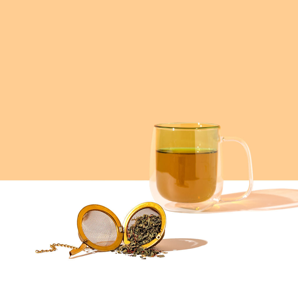 Clear mug with green tea and a gold tea infuser on a white surface with a peach background