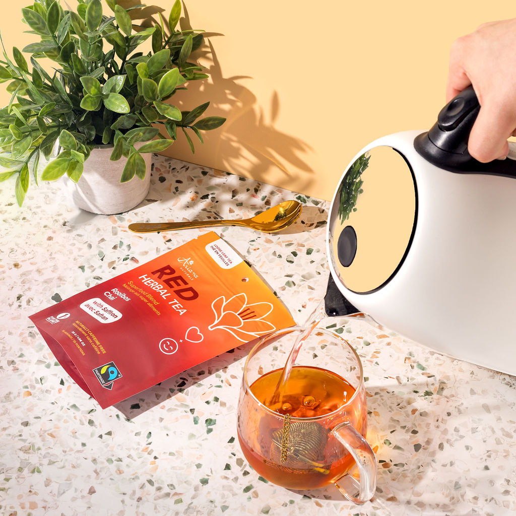 Person pouring tea into a glass mug with a box of Red Tea and a plant on a light surface.