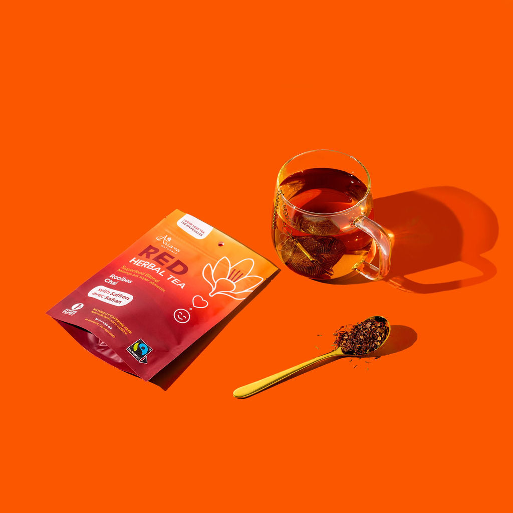 Red herbal tea package, glass mug with tea, and spoonful of tea leaves on an orange background