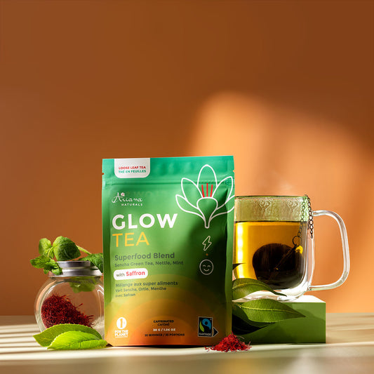 Ariana Naturals Glow Tea package with a mug of the tea and tea ingredients, nettle leaves, sencha green tea leaves, a mint sprig, and a jar of saffron on a green surface and orange background. With afternoon light shining