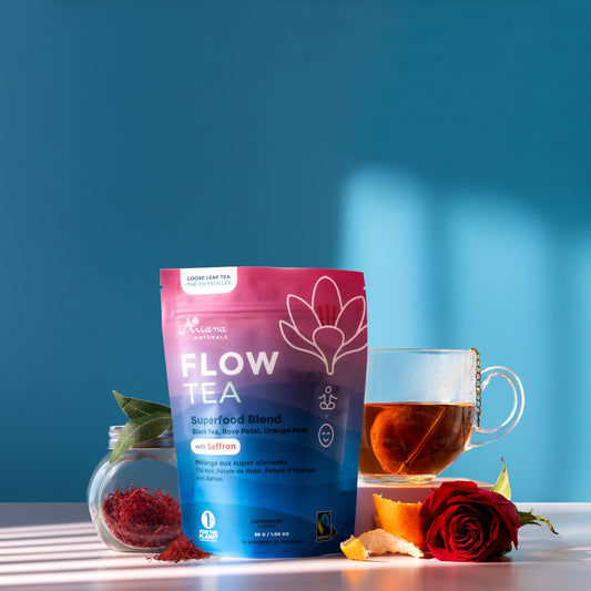 Ariana Naturals Flow Tea package with a cup of the tea and it's ingredients, a jar of saffron, black tea leaves, orange peel twists, and rose on a blue and light pink background. Morning light shining in.