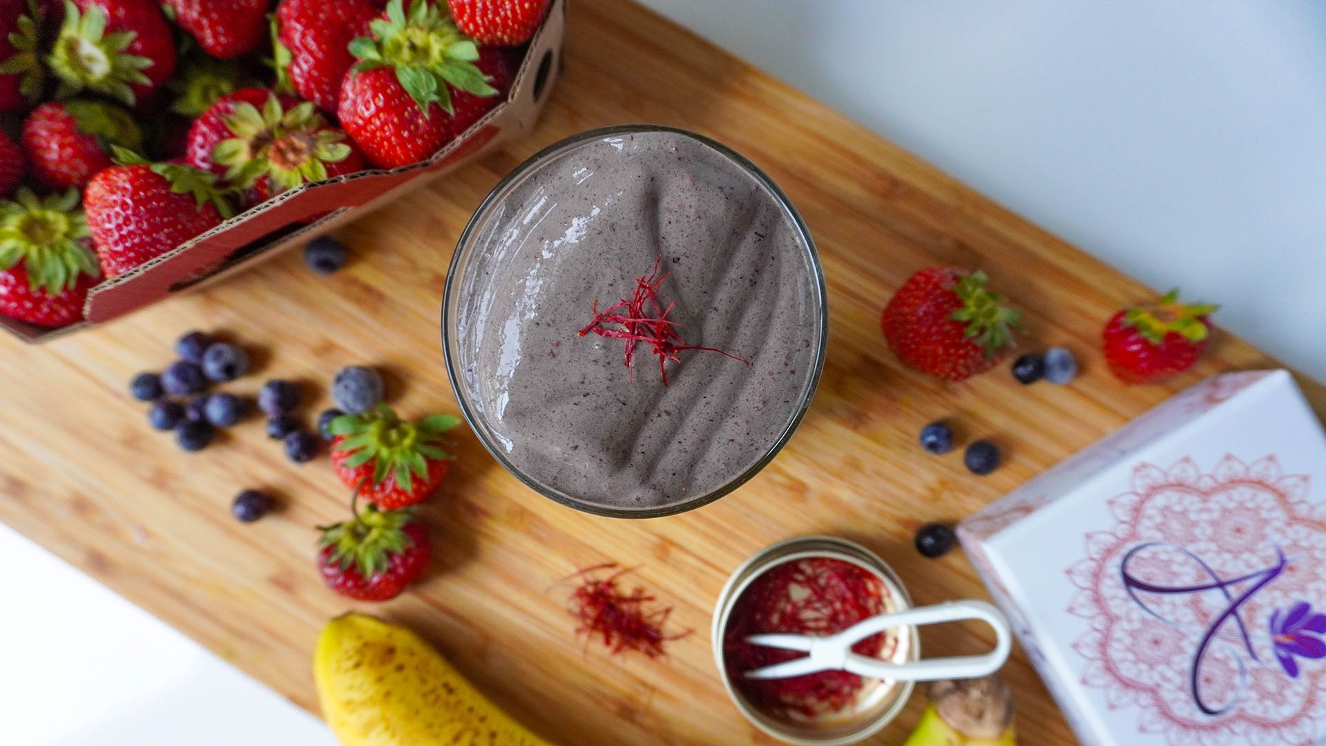 A glass of thick, dark berry smoothie with saffron, styled on a wooden board with fresh strawberries, blueberries, a banana, a small jar of saffron, and a branded Ariana Naturals box.
